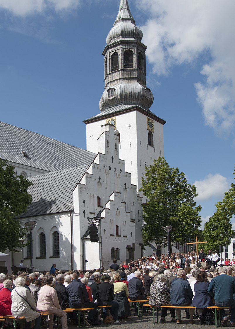 Udendørs gudstjeneste med menighed foran hvid kirke og spir.